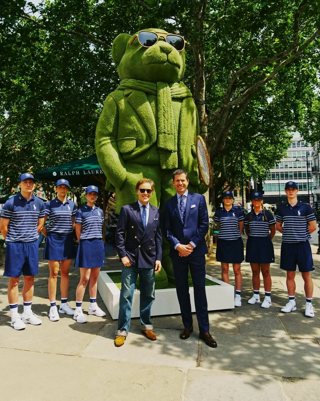 Ralph Lauren Summer of Sport activation in London's Sloane Square: two men in Ralph Lauren tailoring standing in front of a giant green topiary Polo Bear dressed for tennis, with six Polo Ralph Lauren staff in matching navy uniforms flanking them.